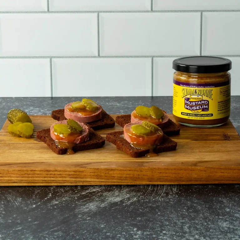 Braunschweiger pumpernickel bites topped with pickle, drizzled with mustard, next to a jar of Slimm & Nunne mustard.