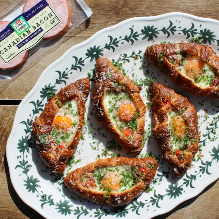 Four egg-topped croissant boats with baked eggs, chives, and parmesan, arranged on a floral platter next to a package of Jones Dairy Farm Canadian Bacon.