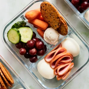 A high protein snack box packed in a divided container with Jones Canadian bacon, hard-boiled eggs, whipped cream cheese, crackers, carrots, cucumber slices, and grapes.