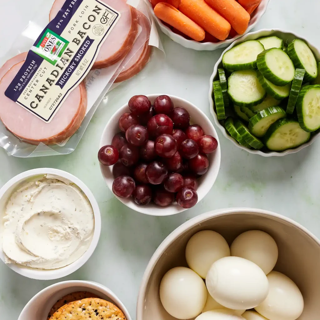 Ingredients for a Canadian bacon protein snack box laid out in bowls, including Jones Hickory Smoked Canadian Bacon, hard-boiled eggs, whipped cream cheese, grapes, cucumber slices, carrots, and crackers.