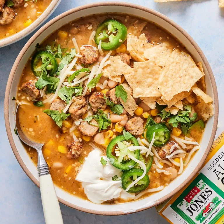 A bowl of creamy white chicken sausage chili with tortilla chips, cheese, jalapeño, sour cream, and cilantro, next to a box of Jones Golden Brown Chicken Sausage.