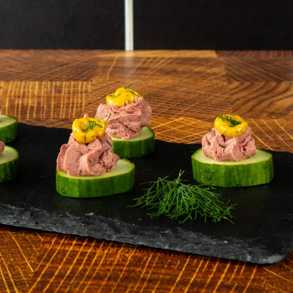 Close-up of cucumber slices topped with whipped Braunschweiger, mustard, and fresh dill on a slate board.
