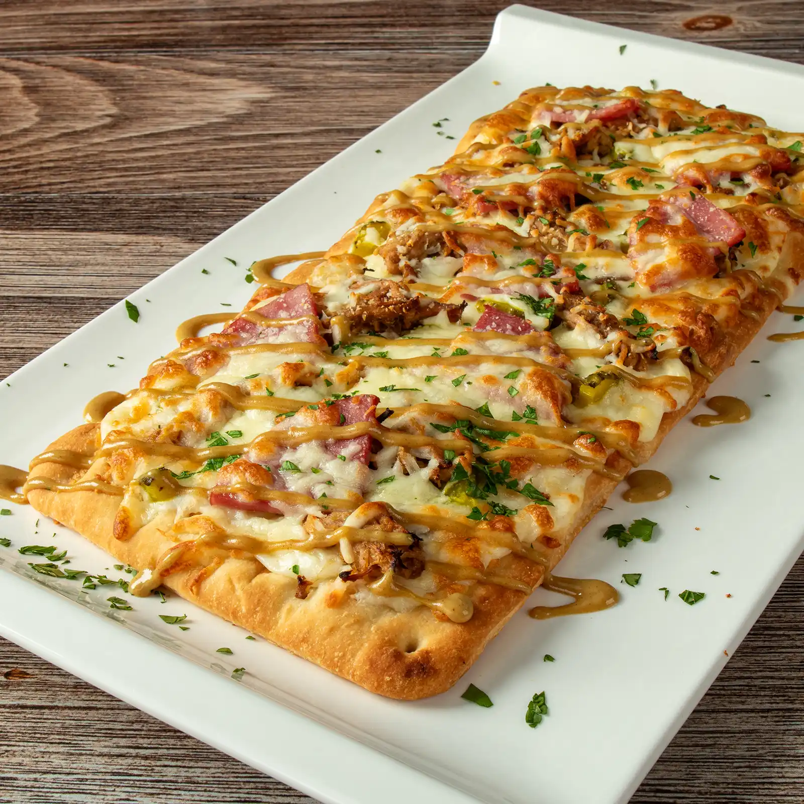 Cubano flatbread with mornay sauce, roast pulled pork, Jones Breakfast Ham, dill pickles, and mozzarella, finished with honey mustard drizzle and parsley