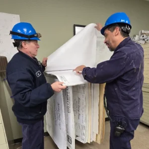 Two Jones Dairy Farm employees in hard hats reviewing large facility blueprints together in a plan room.