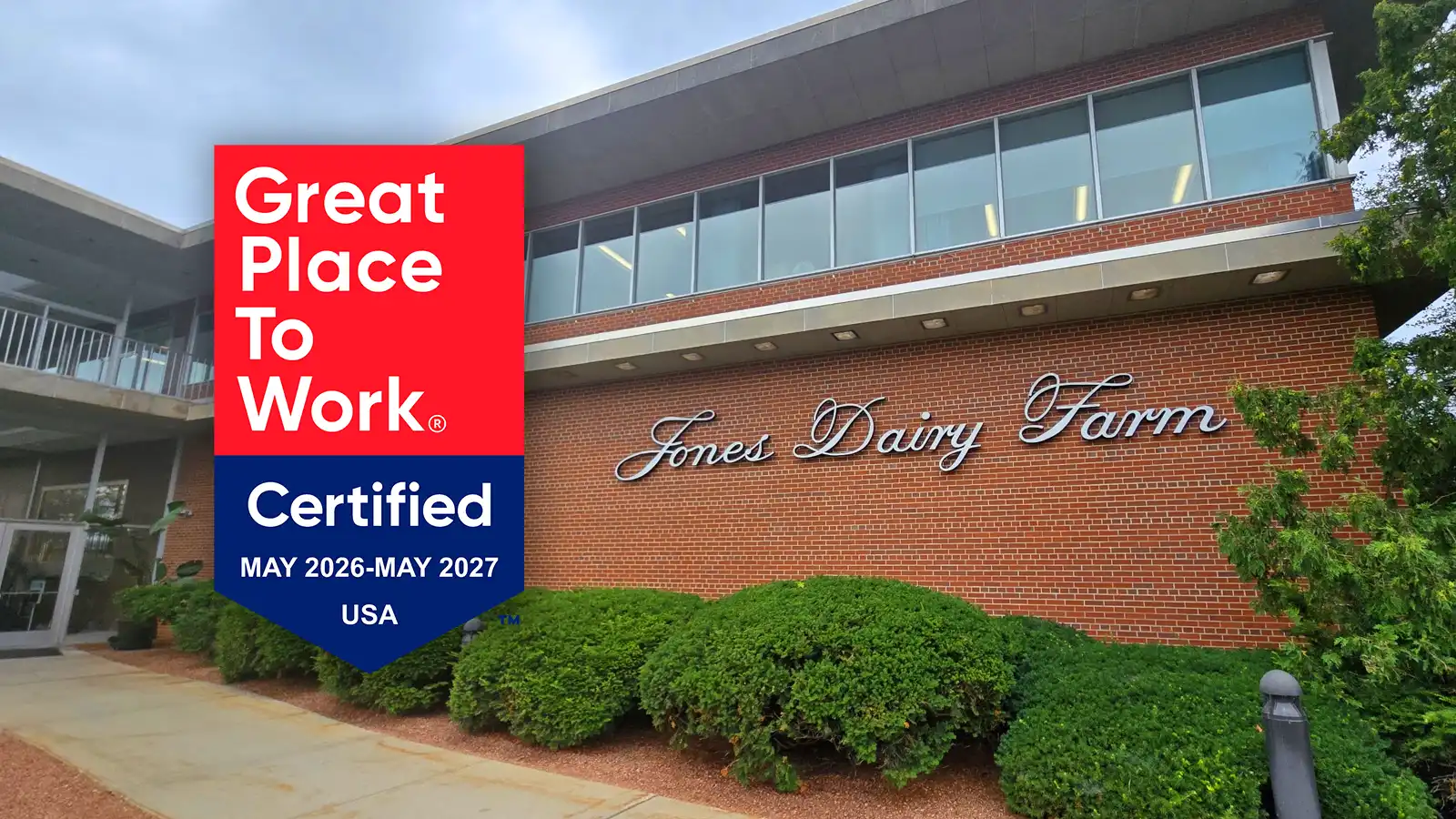 A red and blue Great Place To Work Certified badge for May 2026 to May 2027 overlaid on a photo of the Jones Dairy Farm office building, showing the brick exterior with large windows and the Jones Dairy Farm script logo mounted on the wall above a row of shrubs.