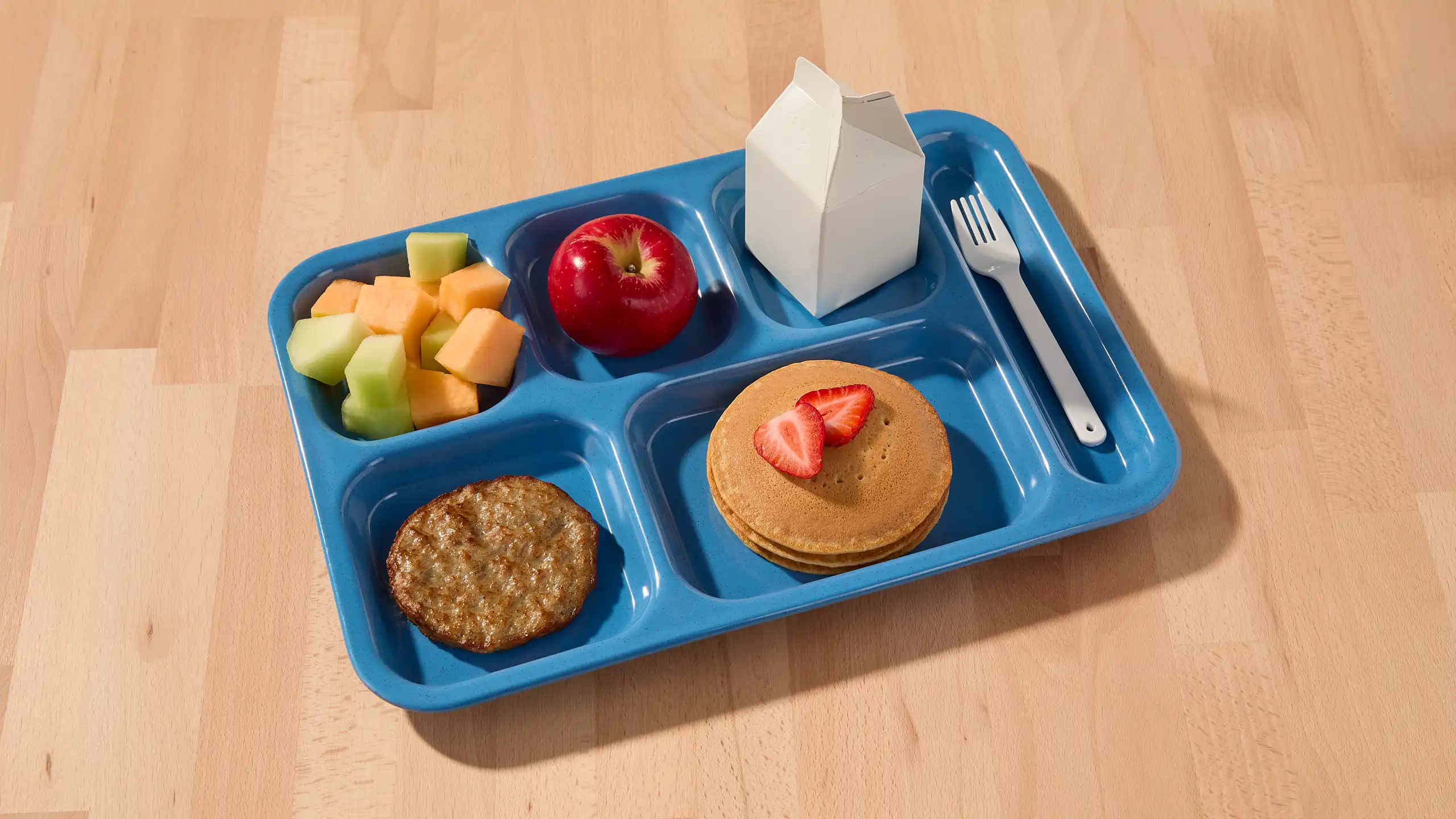 School breakfast tray with a sausage patty, pancakes topped with strawberries, cantaloupe and honeydew, an apple, and a milk carton.