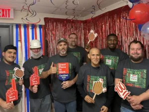A group of seven Jones Dairy Farm production employees celebrate in front of a red tinsel backdrop and streamers, several holding foam hand signs reading 'We're Great' and a giant '100' sign, with one employee holding a Great Place to Work Certified 2025 award, all wearing the same gray People-First, Family-Owned T-shirts.