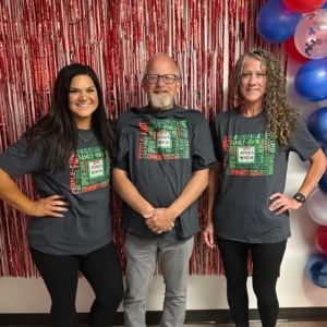 Three Jones Dairy Farm employees stand smiling in front of a red tinsel backdrop with red, white, and blue balloons, each wearing a gray T-shirt printed with words like People-First, Family-Owned, and Employee-Centered around the Jones Dairy Farm logo, celebrating the company's Great Place to Work certification.