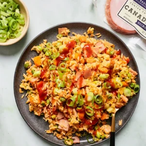 Overhead view of a plate of kimchi fried rice with Canadian bacon, topped with green onion and sriracha, next to a bowl of sliced green onion and a package of Jones Canadian bacon.