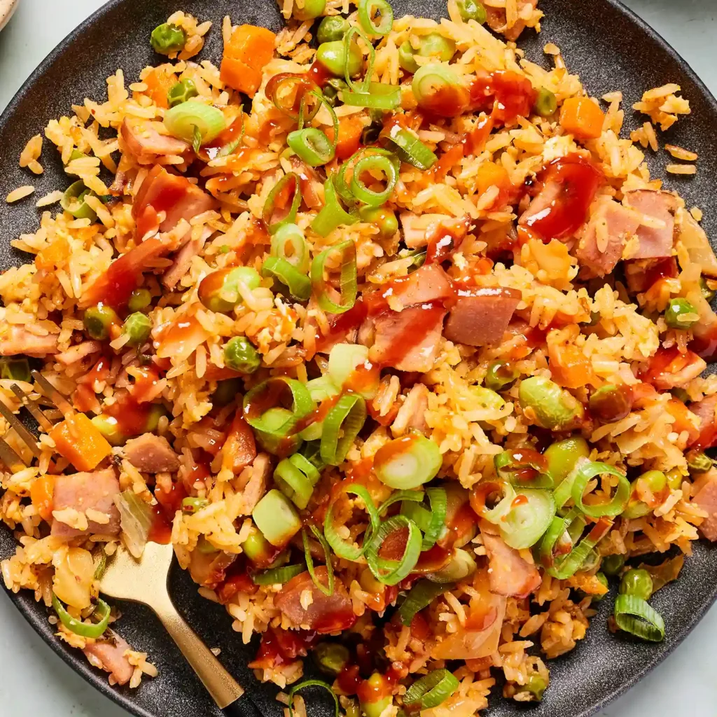 Close-up of kimchi fried rice with Canadian bacon, peas, edamame, carrots, and green onion, drizzled with sriracha.