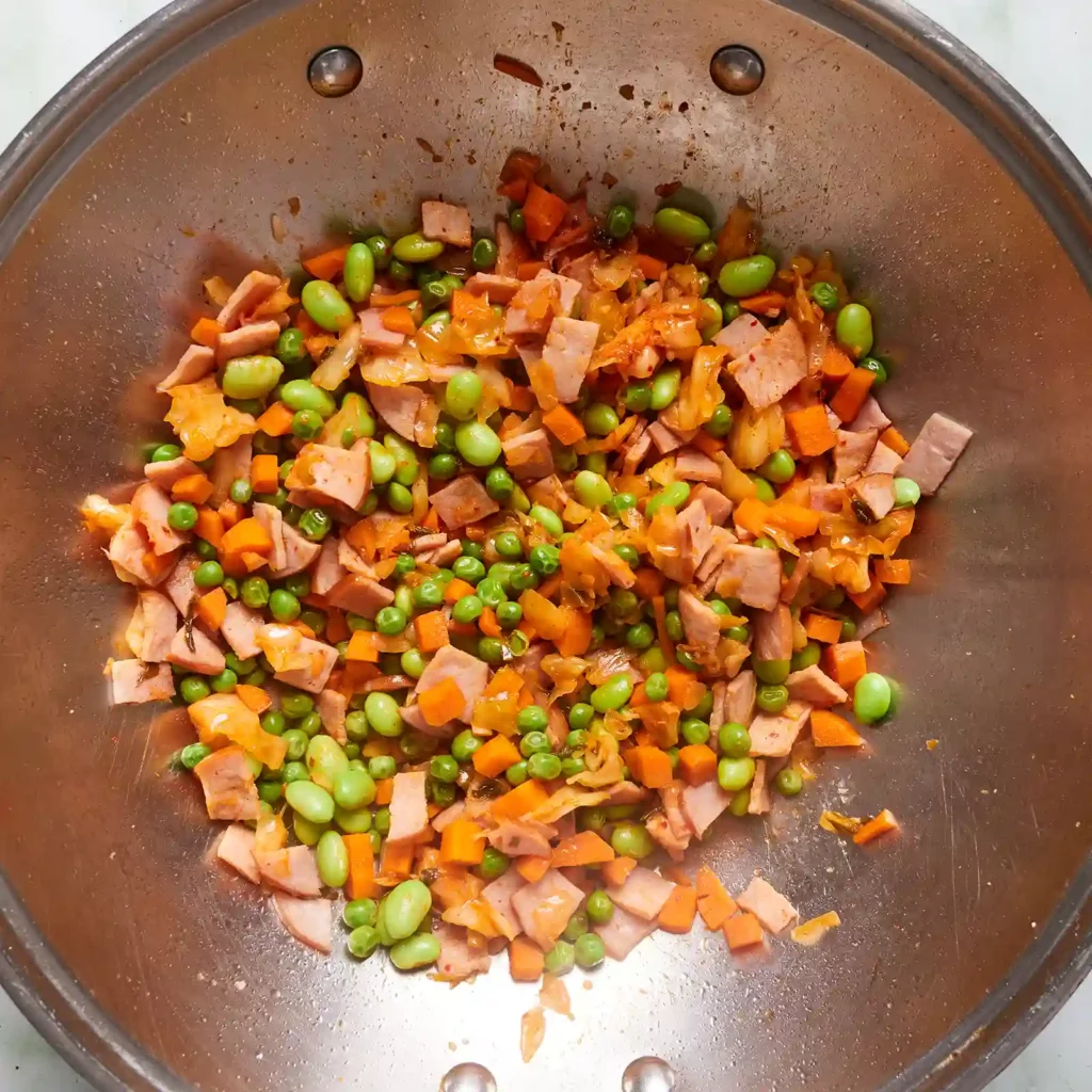 Diced Canadian bacon, carrots, peas, edamame, and kimchi cooking together in a wok for kimchi fried rice.