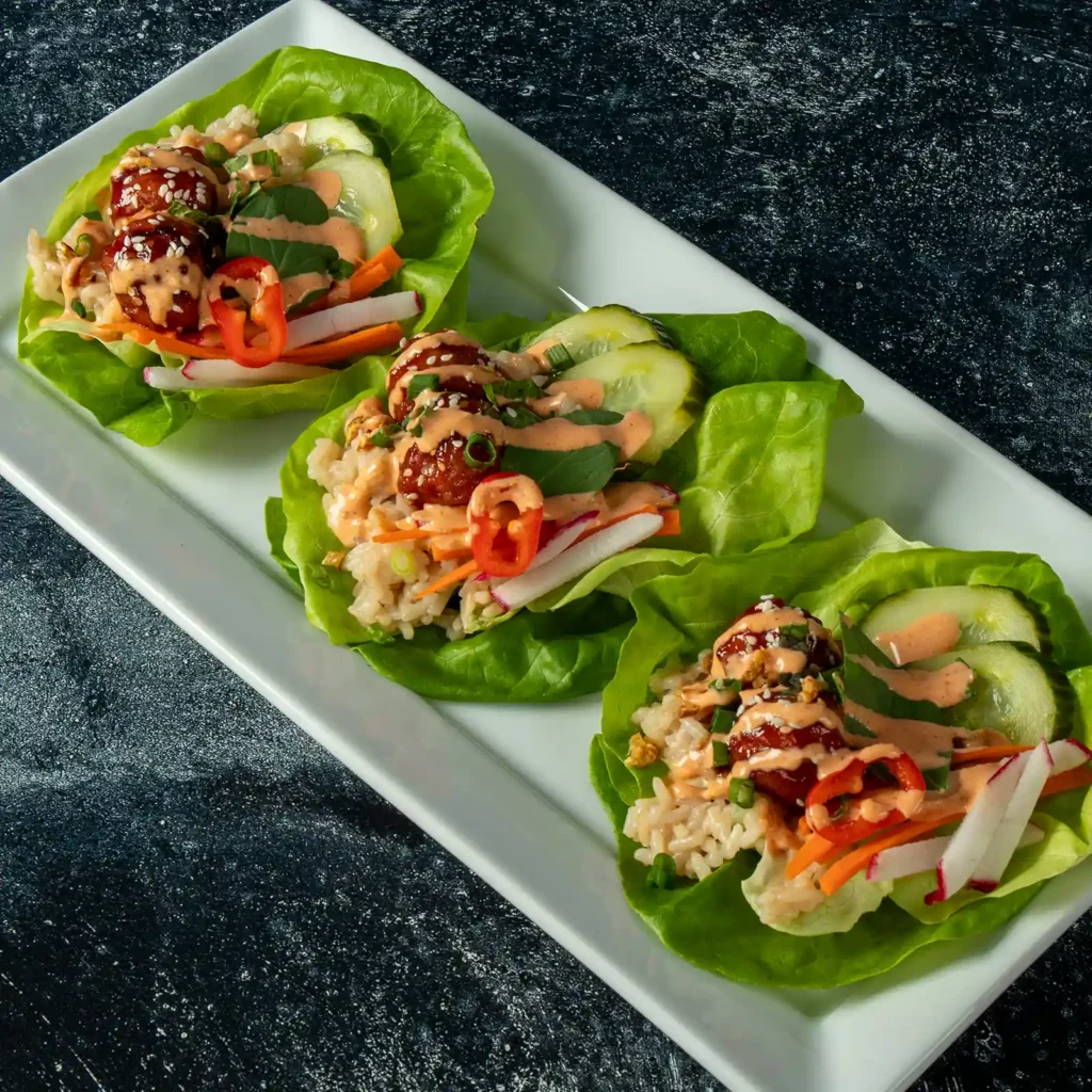 Overhead view of three Korean BBQ lettuce wraps with Jones Pork and Bacon Meatballs, sushi rice, radish, carrots, and cucumber