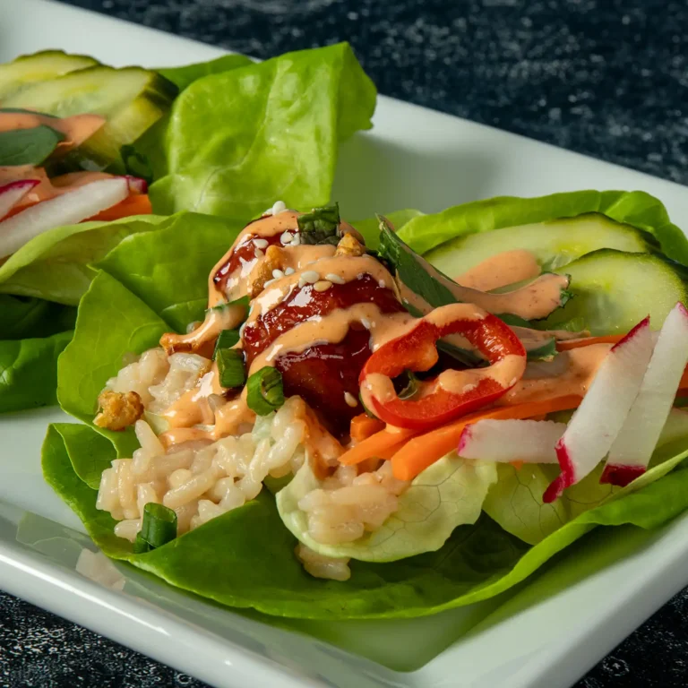 Close-up of a Korean BBQ lettuce wrap showing glazed meatball, sesame seeds, Fresno chili, and sriracha mayo drizzle