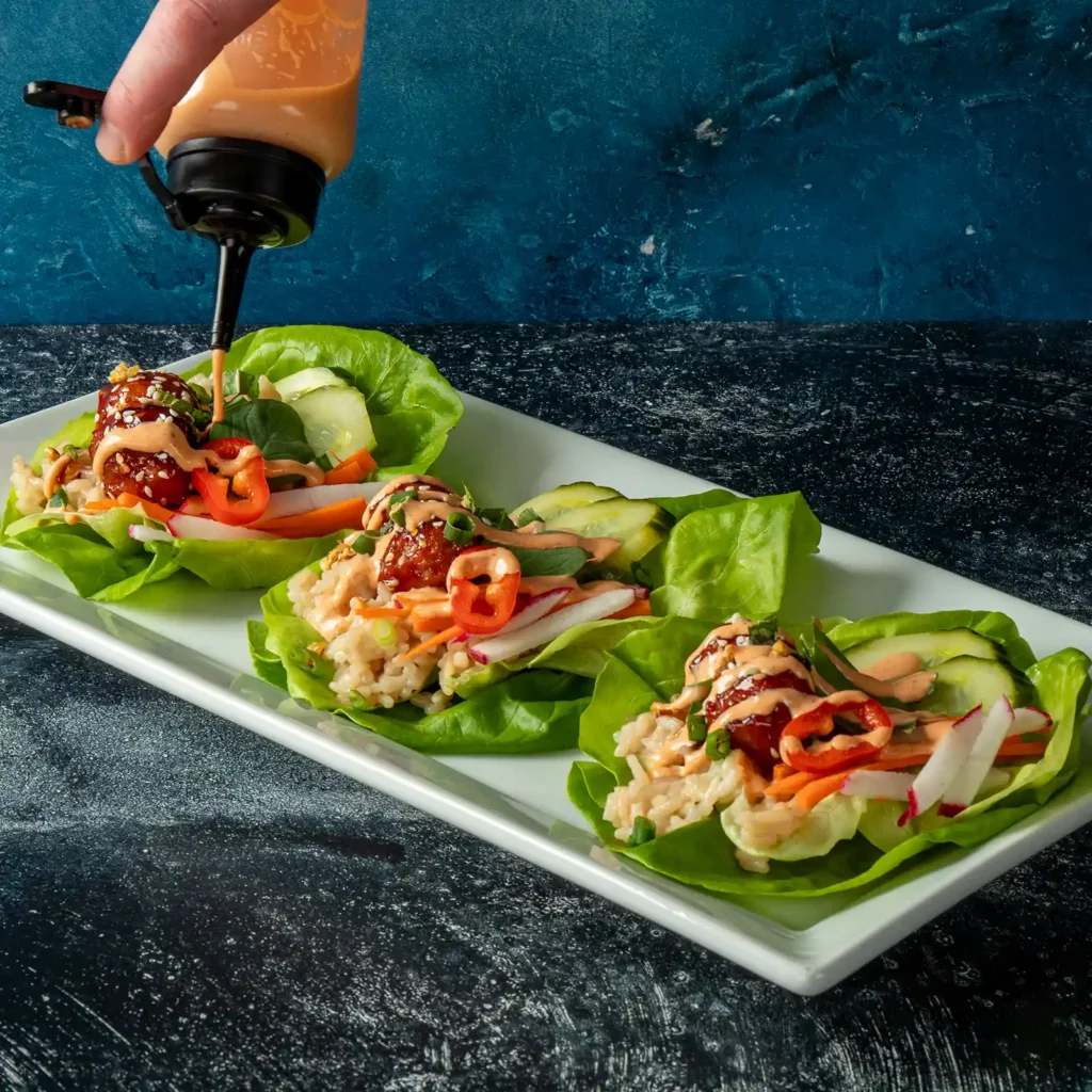 Close-up of sriracha mayo being poured over a Korean BBQ meatball lettuce wrap on a white rectangular plate