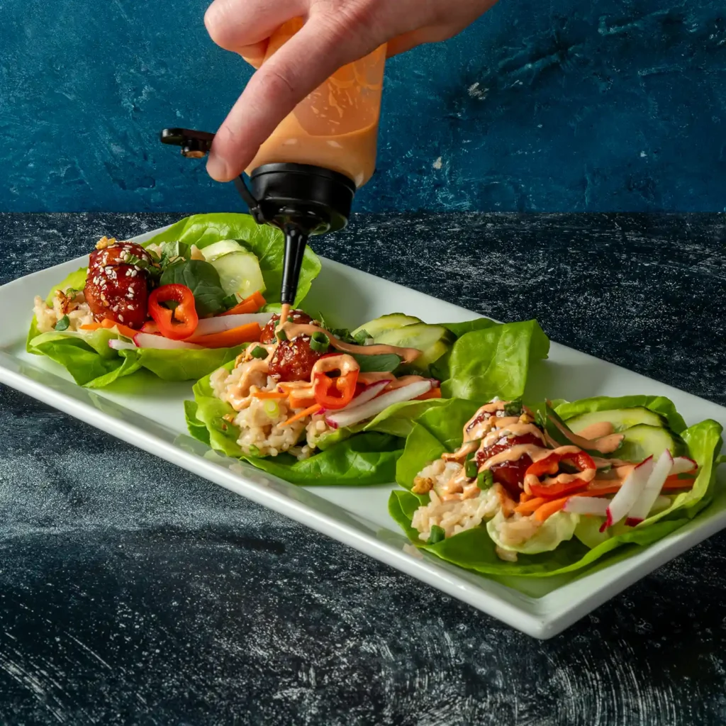 Hand drizzling sriracha mayo over Korean BBQ lettuce wraps with Jones Pork and Bacon Meatballs, sushi rice, and fresh vegetables
