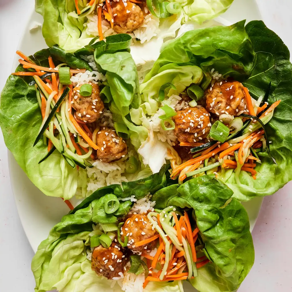 Overhead view of several lettuce wraps with soy sesame chicken meatballs, rice, pickled cucumber and carrot, green onion, and sesame seeds, arranged on a platter.