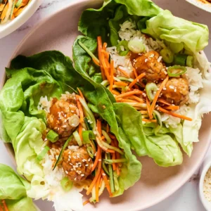 Two lettuce wraps filled with rice, soy sesame chicken meatballs, quick-pickled cucumber and carrot, green onion, and sesame seeds.
