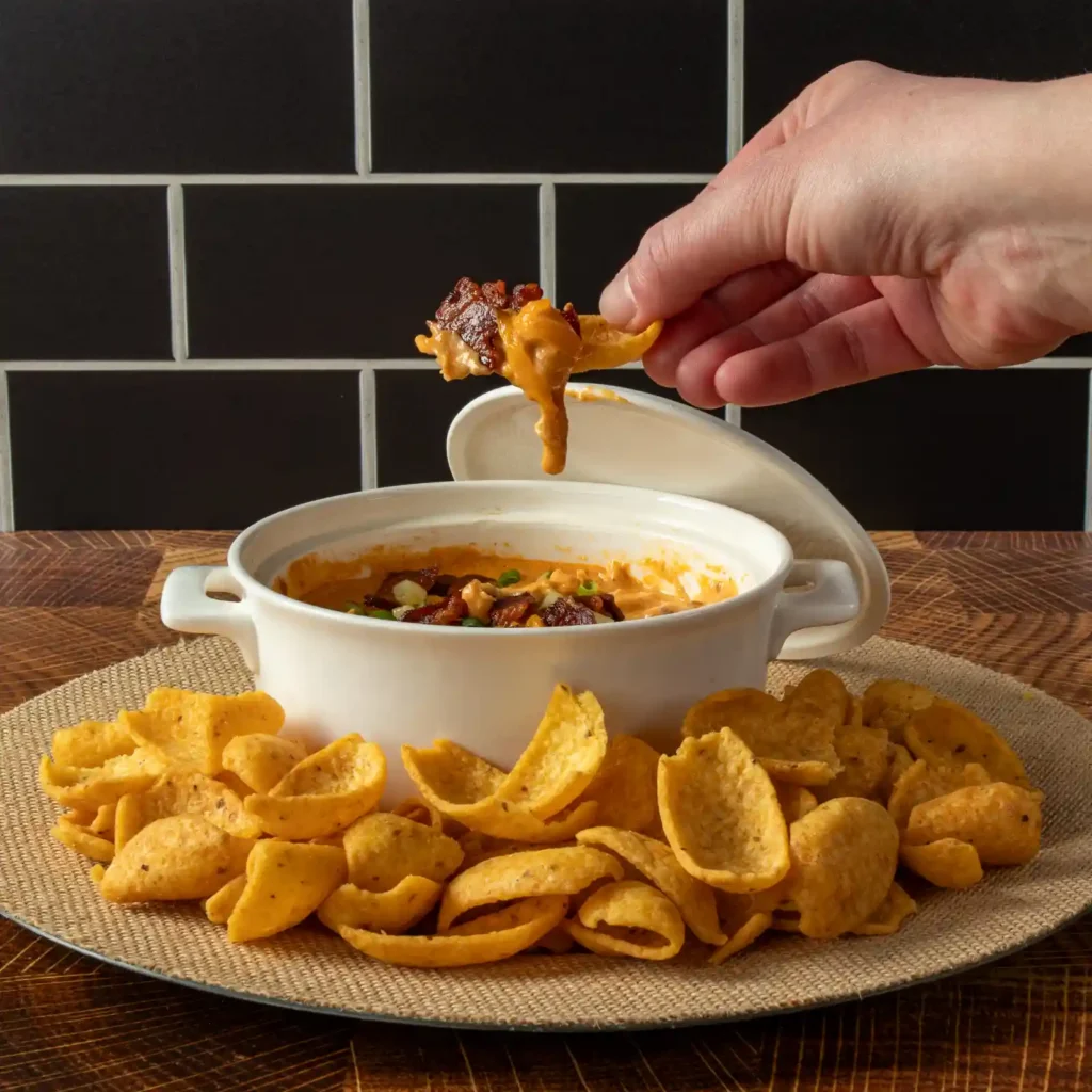 A hand dips a corn chip into a bowl of Nashville hot chicken dip topped with bacon, scooping up a bite loaded with cheese and crispy bacon bits.
