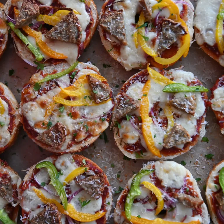 Overhead view of a tray of sausage and pepper pizza bagels, each topped with marinara, melted cheese, pepper strips, red onion, and grated parmesan.