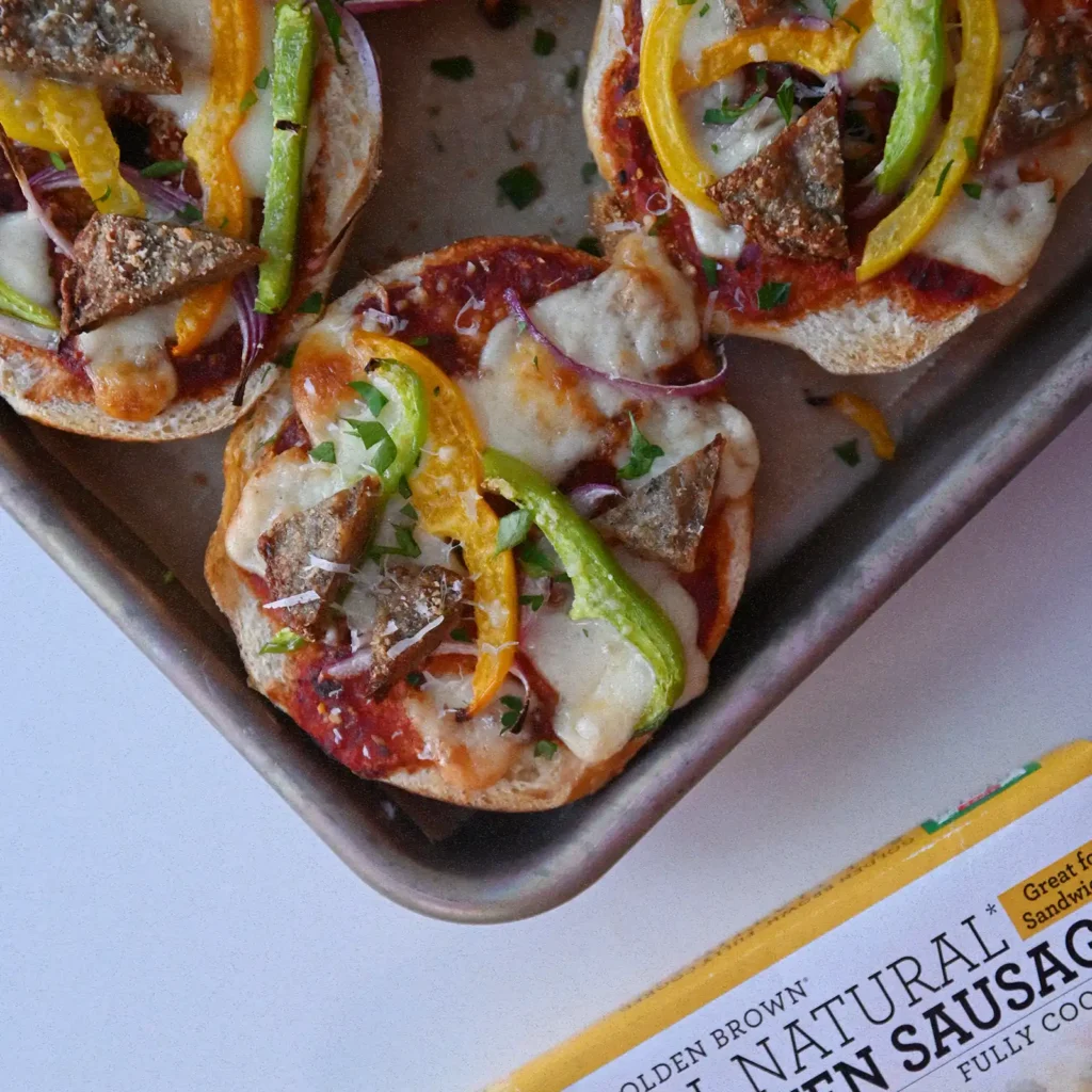 Sausage and pepper pizza bagels topped with melted mozzarella, yellow and green peppers, red onion, and parsley on a baking sheet, next to a box of Jones Golden Brown Chicken Sausage.