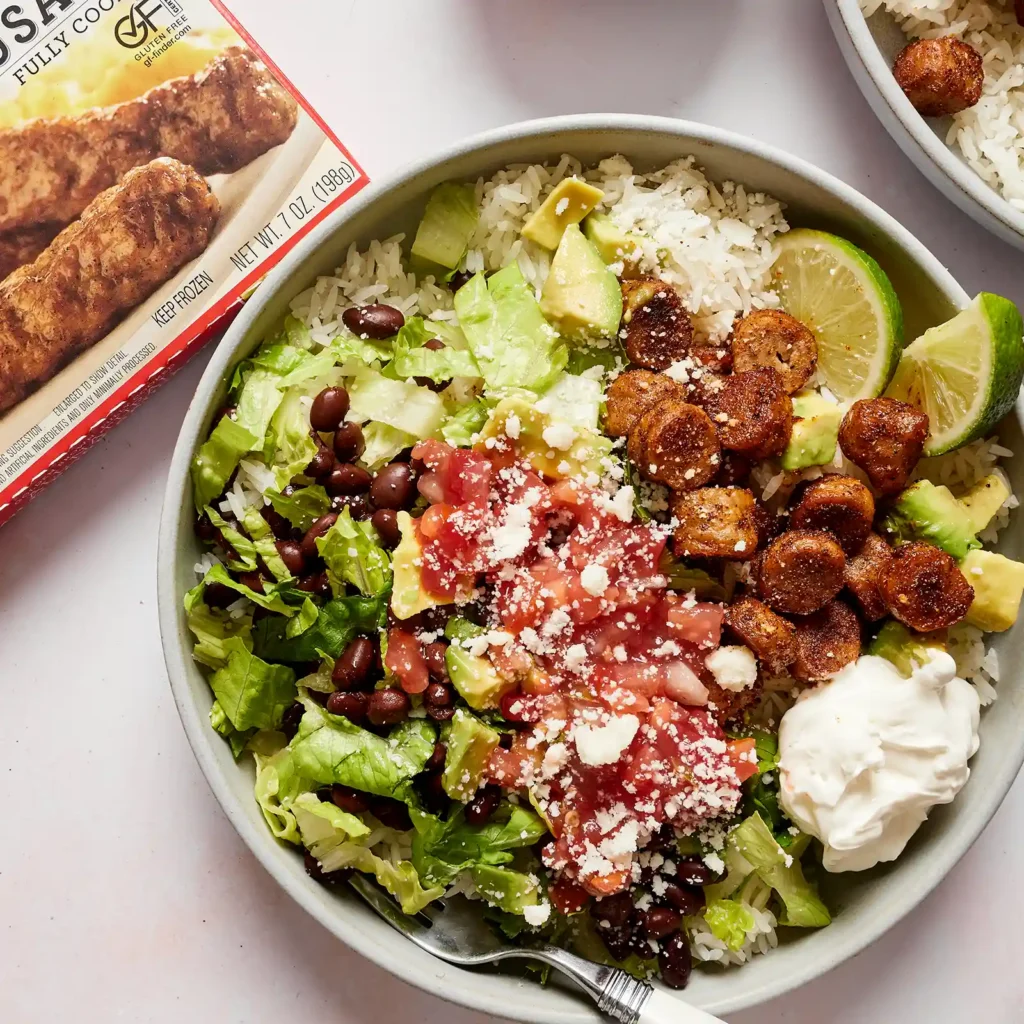 A taco rice bowl with sliced Jones sausage, rice, black beans, lettuce, avocado, pico de gallo, cotija cheese, sour cream, and lime, next to the Jones sausage box.