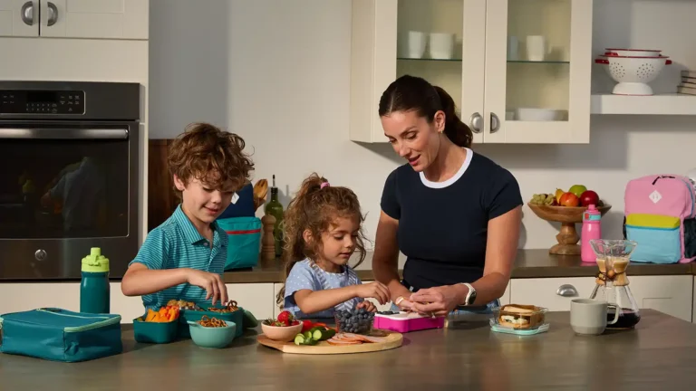 Mom packing school lunches with her son and daughter at the kitchen counter, filling containers with grapes, carrots, pretzels, and sandwich fixings.