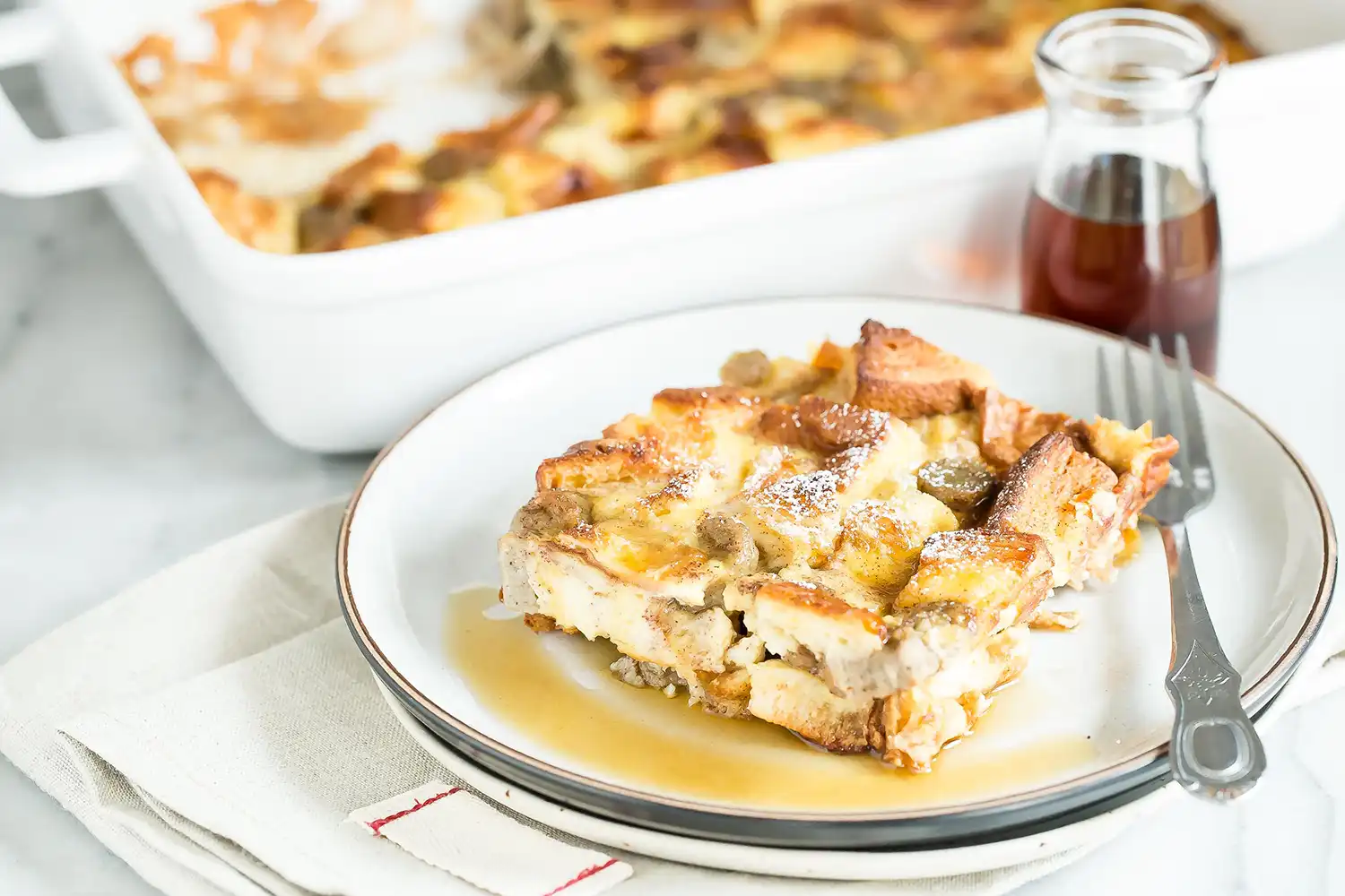 Slice of cinnamon French toast bake with sausage on a plate, drizzled with maple syrup, with the full casserole dish and a syrup bottle in the background.