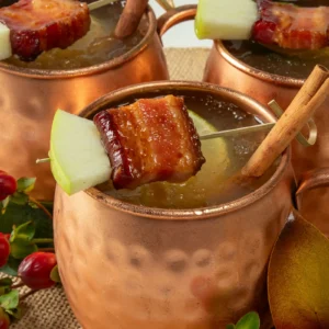 Bacon Apple Mule cocktails in copper mugs, garnished with a skewer of cinnamon-glazed Jones Dry Aged Cherrywood Smoked Slab Bacon, apple slice, and a cinnamon stick