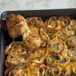 Tray of baked Mexican sausage pinwheels with melted golden cheese, one being lifted with tongs showing a cheese pull