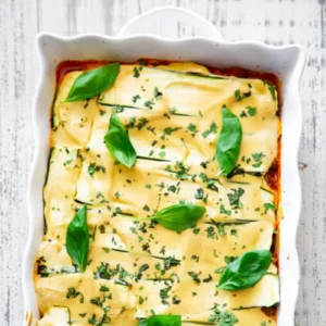 Overhead view of zucchini lasagna in a white baking dish, with melted cashew cheese sauce, chopped herbs, and fresh basil leaves on top.
