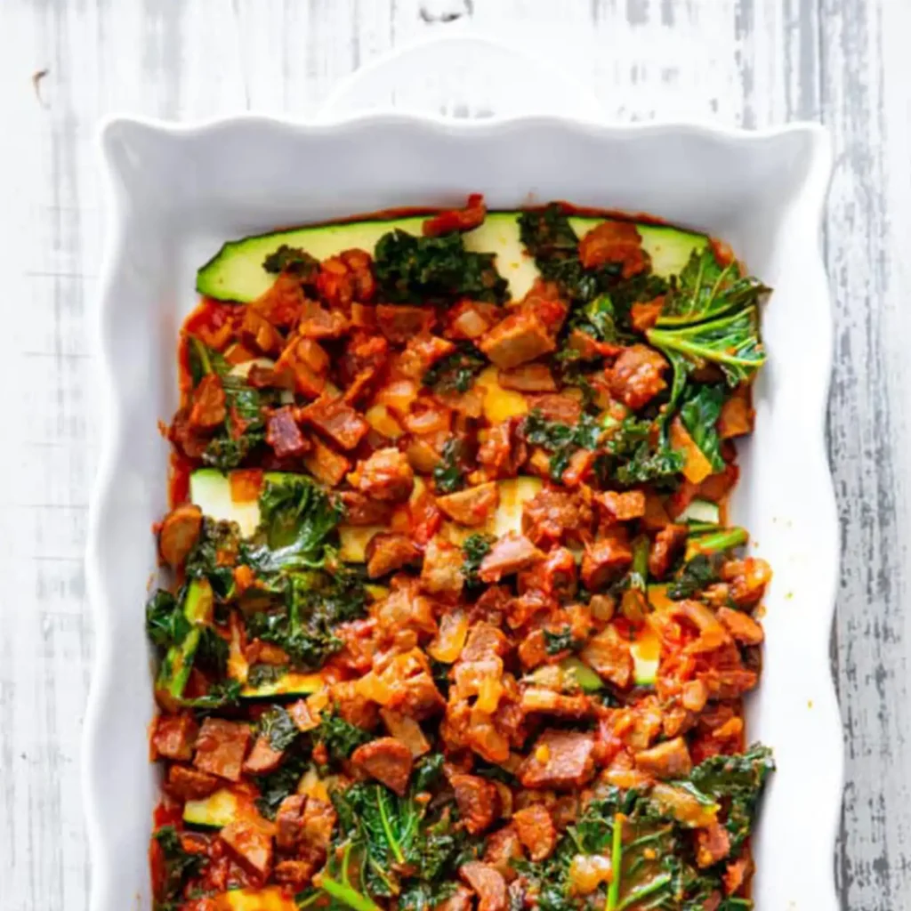 Zucchini lasagna being layered in a white baking dish, with a chopped sausage, kale, and tomato sauce mixture spread over zucchini strips.