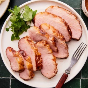 Sliced apricot Dijon glazed ham on a plate garnished with parsley, with a bowl of dipping sauce alongside.