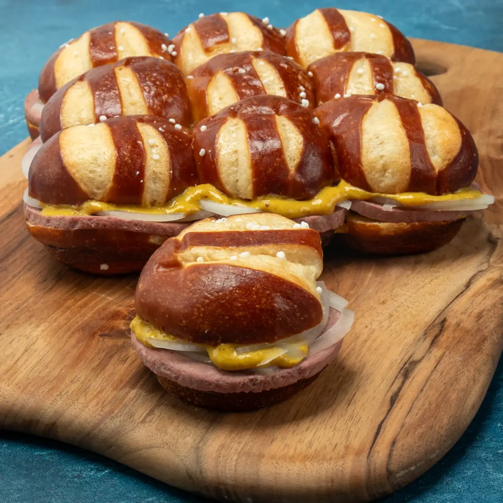 Braunschweiger slider sandwiches on pretzel buns with mustard and onion, arranged on a wood board.