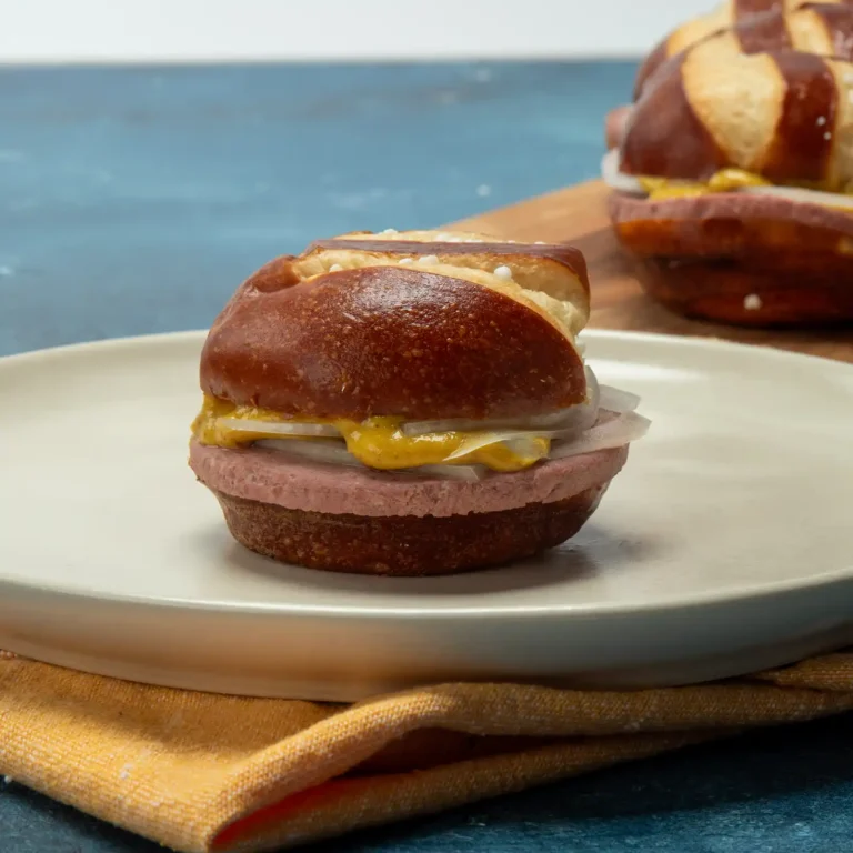 Braunschweiger slider sandwich on a pretzel bun with mustard and onion, plated with more sliders in the background.