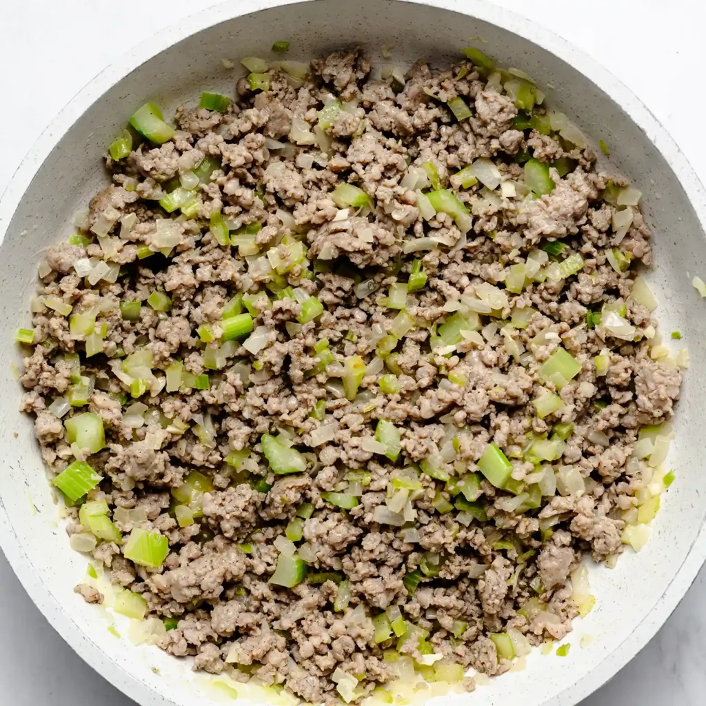 Ground Jones sausage browning with celery and onion in a skillet, the base for sourdough sausage stuffing.