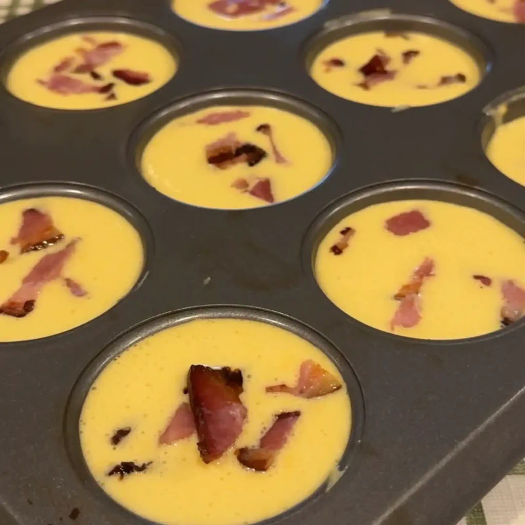 Egg mixture poured over baked tater tots and bacon pieces in a muffin tin, ready to bake.