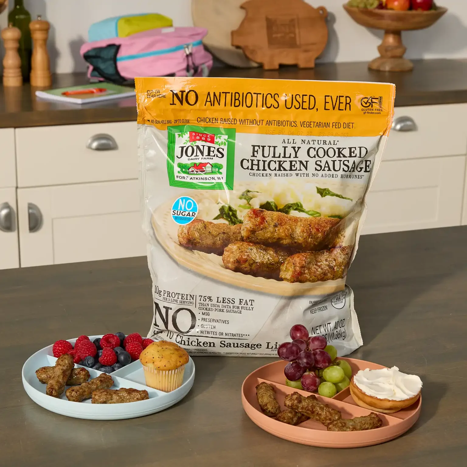 Jones Dairy Farm Fully Cooked Chicken Sausage bulk bag on a kitchen counter beside two kids' divided plates with sausage links, berries, grapes, a muffin, and a bagel with cream cheese.