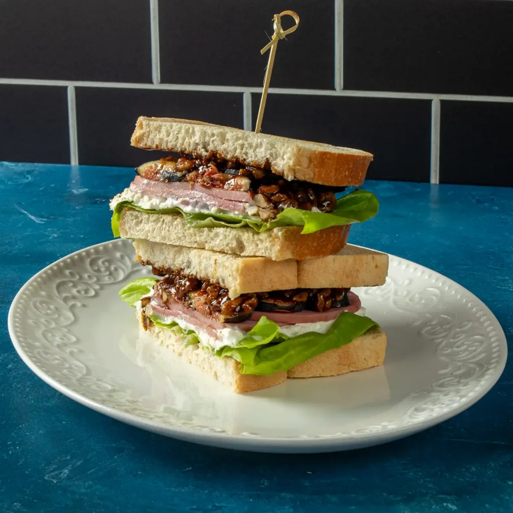 Two halves of a fig and ham sandwich stacked on a plate, layered with whipped feta, ham, fig and walnut topping, and gem lettuce between slices of white bread.