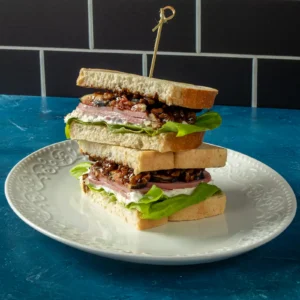Two halves of a fig and ham sandwich stacked on a plate, layered with whipped feta, ham, fig and walnut topping, and gem lettuce between slices of white bread.