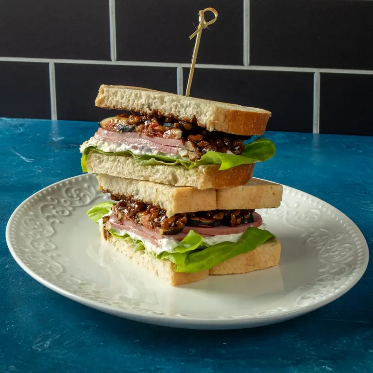 Two halves of a fig and ham sandwich stacked on a plate, layered with whipped feta, ham, fig and walnut topping, and gem lettuce between slices of white bread.