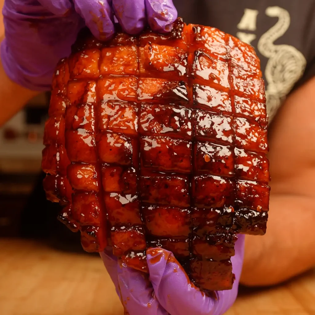 Close-up of a glazed ham slice held in a gloved hand, showing the deep mahogany color and diamond-scored crust from the hot honey chipotle bourbon glaze.