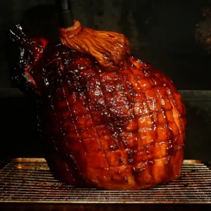 Whole hickory smoked ham with a dark, caramelized hot honey chipotle bourbon glaze, scored in a diamond pattern, resting on a wire rack in a smoker.
