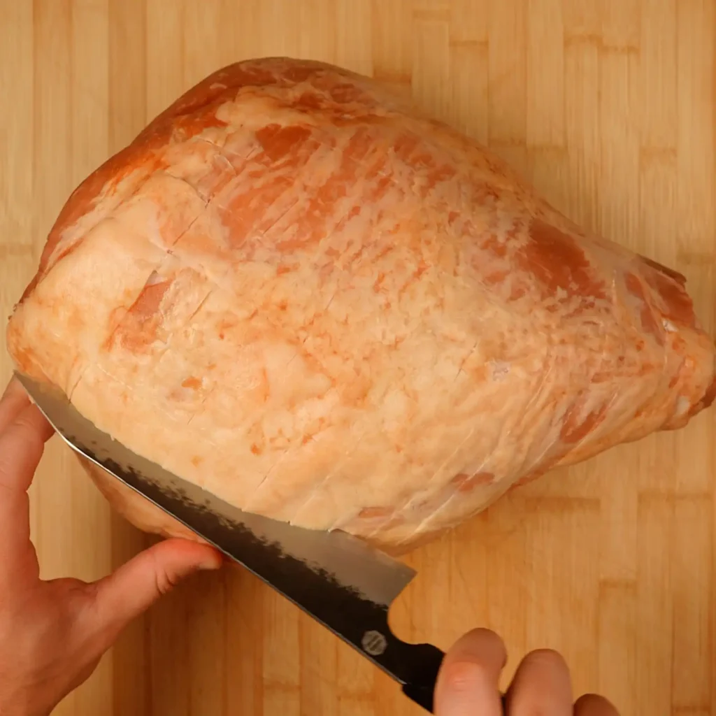 Raw whole ham scored in a diamond pattern on a wood cutting board, with a knife mid-cut along the scored lines.