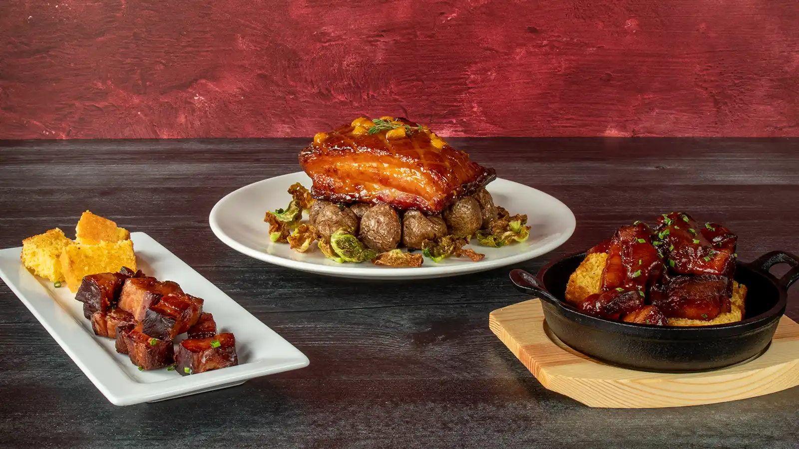 Three plated dishes featuring glazed dry aged bacon slab: diced bacon cubes with cornbread on a white plate, a whole glazed bacon slab over roasted potatoes and brussels sprout leaves, and bacon cubes in a cast iron skillet with cornbread.