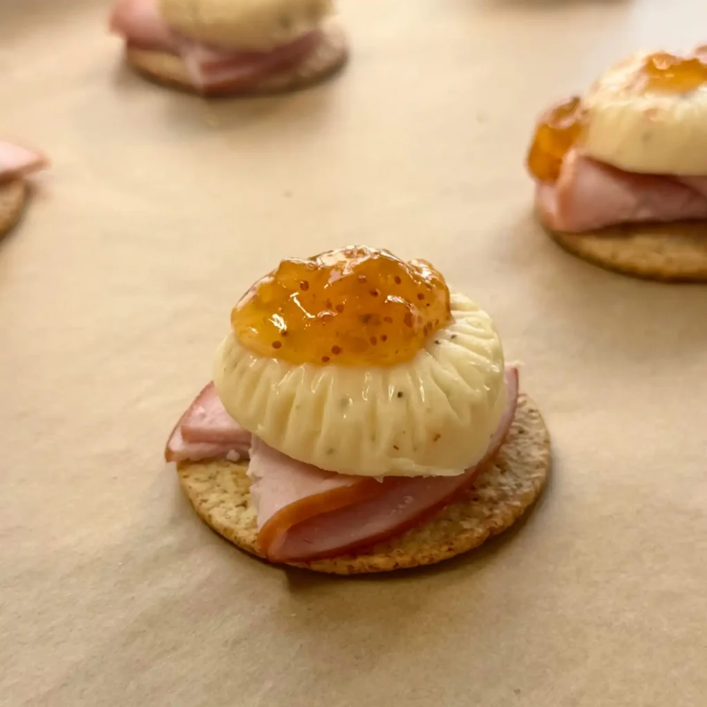 Cracker topped with Canadian bacon, a round of cheese, and fig jam before broiling, with more assembled bites in the background.