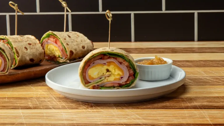 Sliced breakfast wrap with scrambled egg, ham, cheese, tomato, and lettuce, served on a plate with a side of apple butter, additional wraps on a wooden board in the background.
