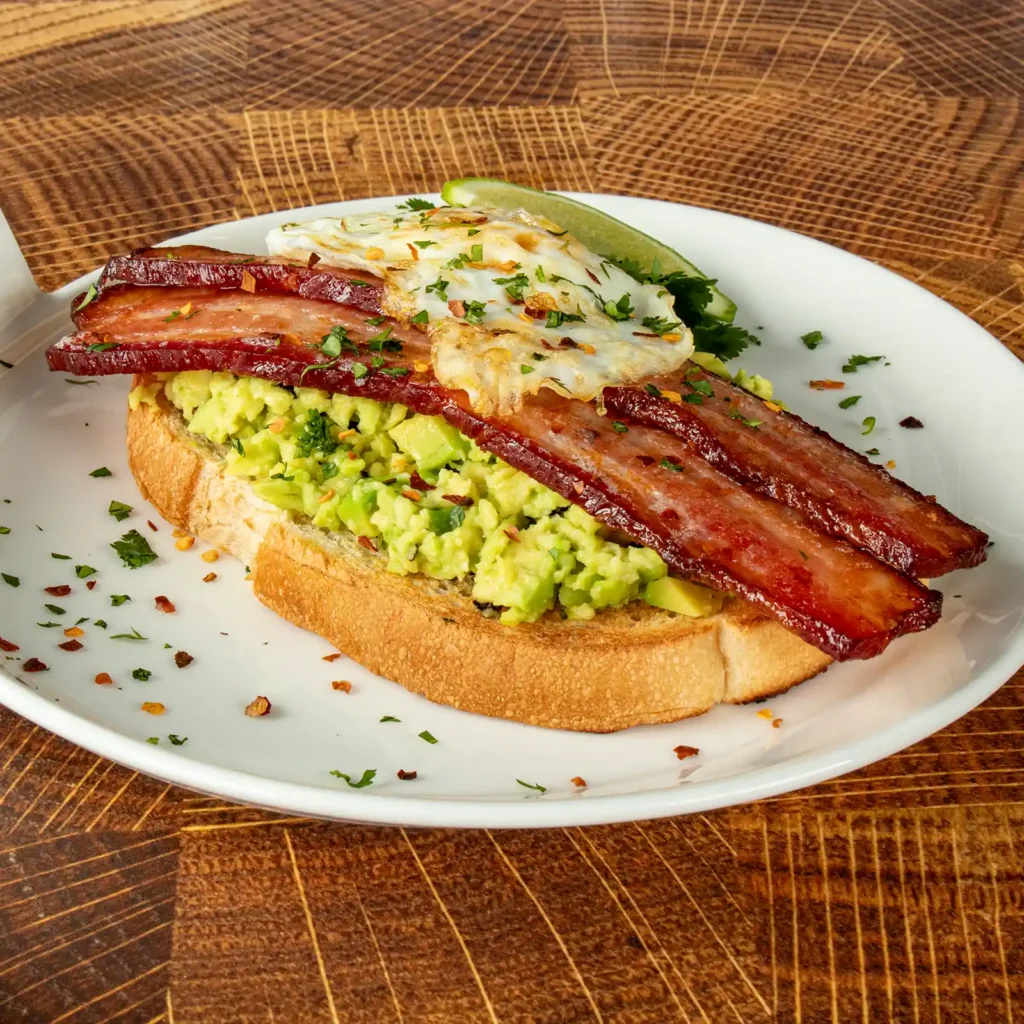 Thick-cut seared bacon slices over smashed avocado toast with a fried egg, chili flakes, and fresh herbs