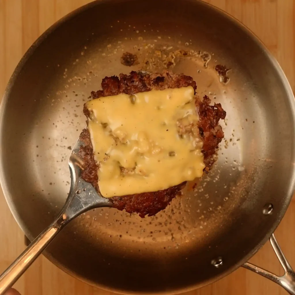 Seared beef patty topped with melted American cheese on a spatula over a stainless steel skillet