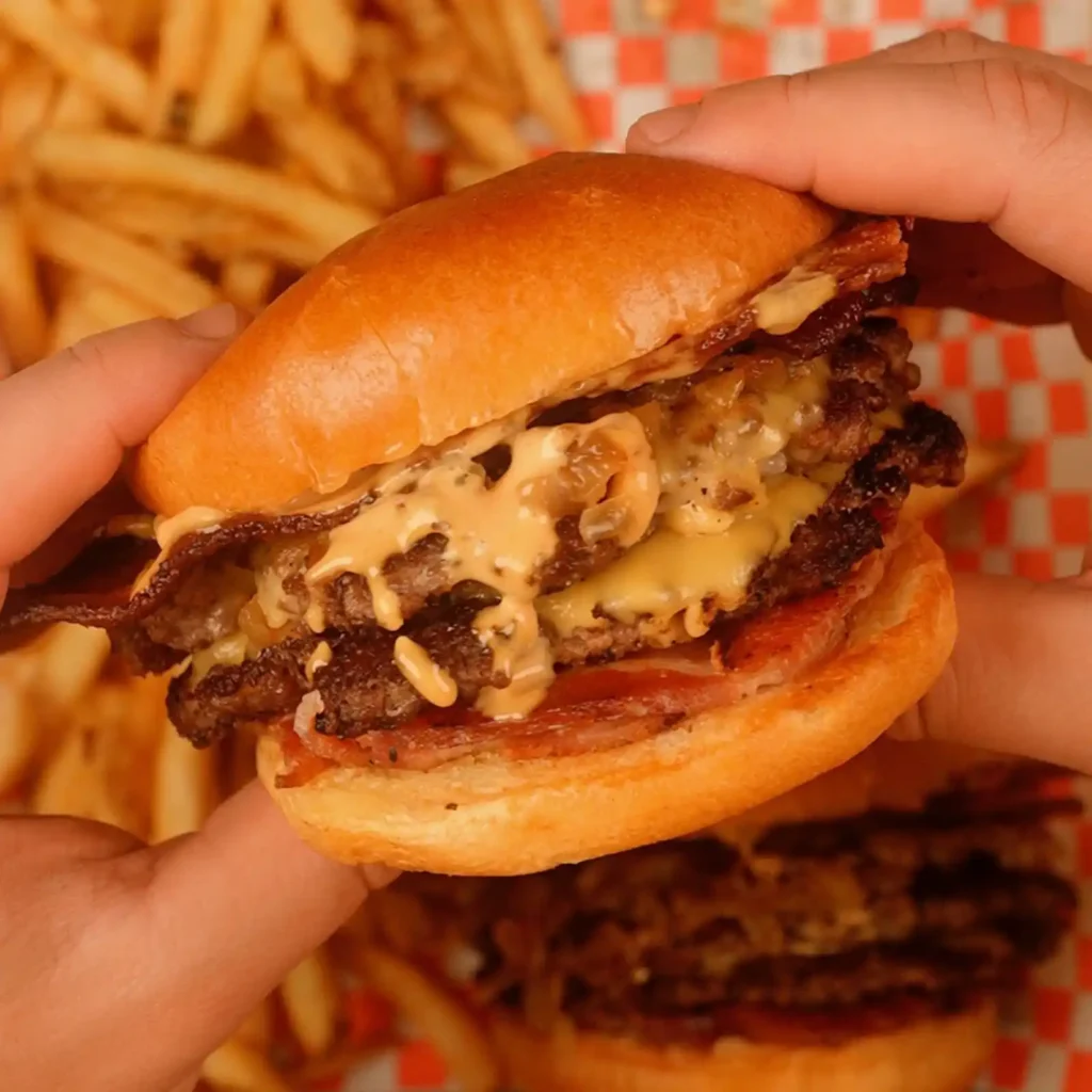 Hands holding a bacon cheeseburger with two patties, melted cheese, caramelized onions, and burger sauce dripping from a brioche bun