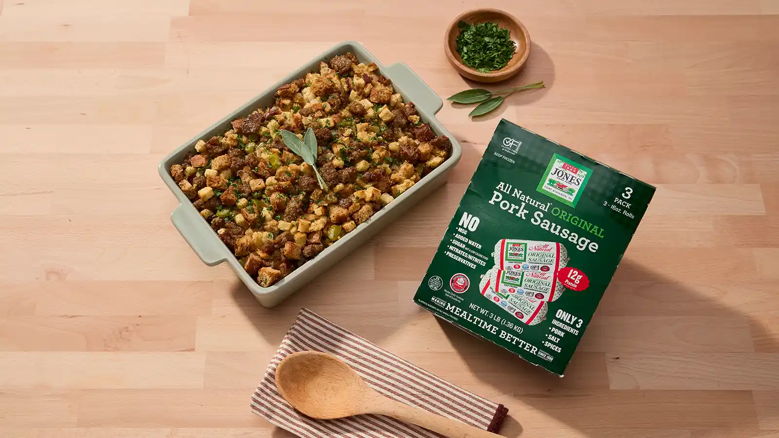 Sausage stuffing in a sage green baking dish, garnished with a fresh sage sprig, beside a 3-pack box of Jones All Natural Original Pork Sausage on a wooden counter.